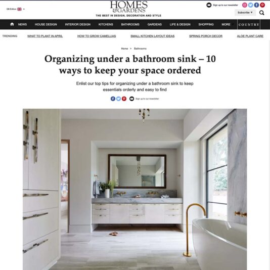 A screenshot of a *Homes & Gardens* article titled "Organizing under a bathroom sink – 10 ways to keep your space ordered." The page features a banner menu, a header image of a bathroom with a wooden vanity, sinks, and a large mirror, and the article text below.