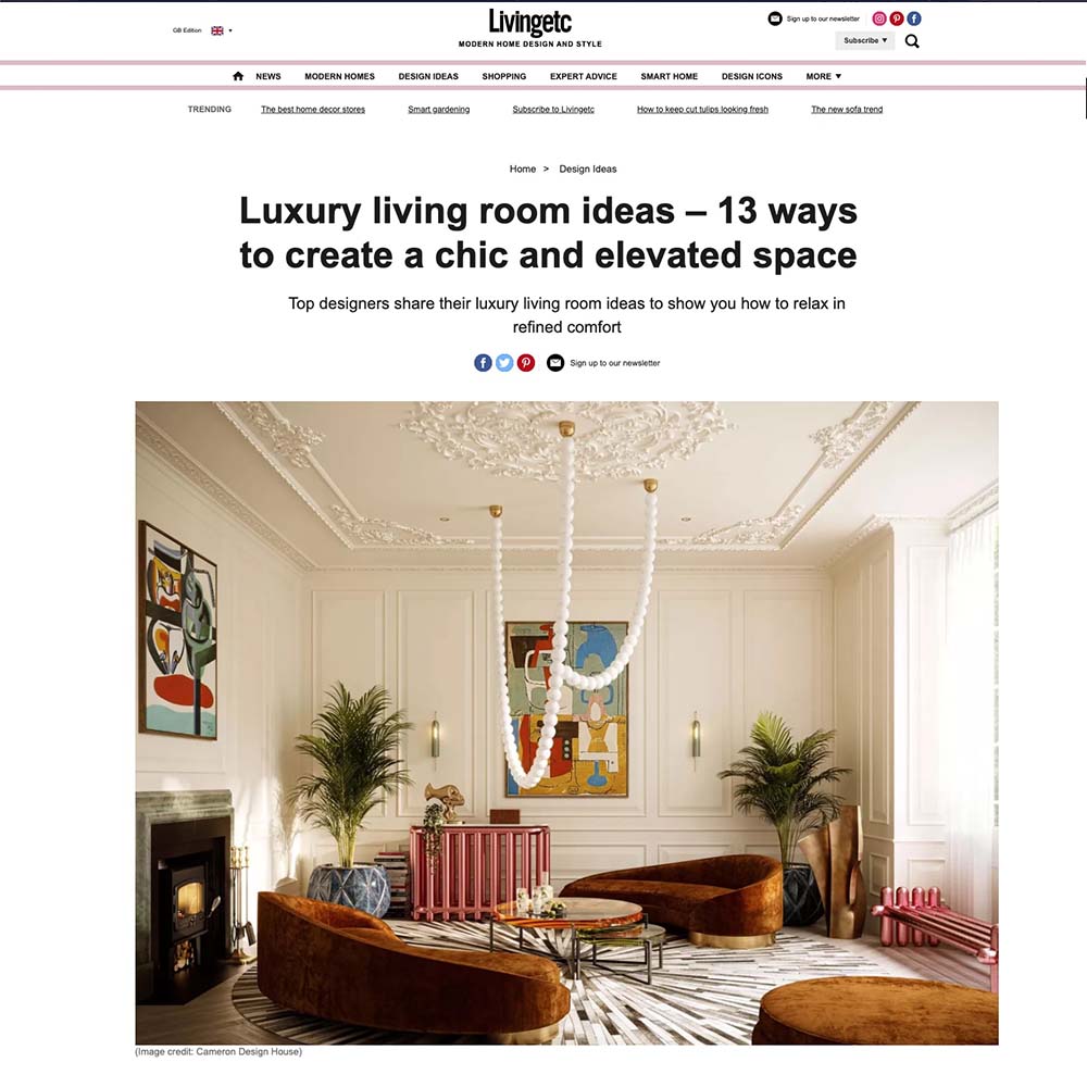 A luxurious living room with ornate white ceilings and a neutral color palette. The room features brown velvet sofas, a circular gold-trimmed coffee table, and contemporary artwork on the walls. A large chandelier hangs from the ceiling, adding an elegant touch.