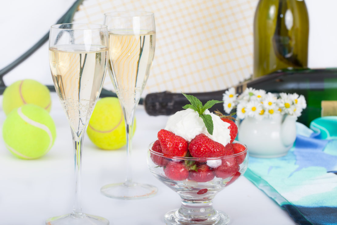 A Wimbledon-style lawn party setup features two champagne glasses, a bowl of strawberries with whipped cream and mint, tennis balls, a tennis racket, a wine bottle, and a small vase with daisies on a table adorned with a blue and white cloth.