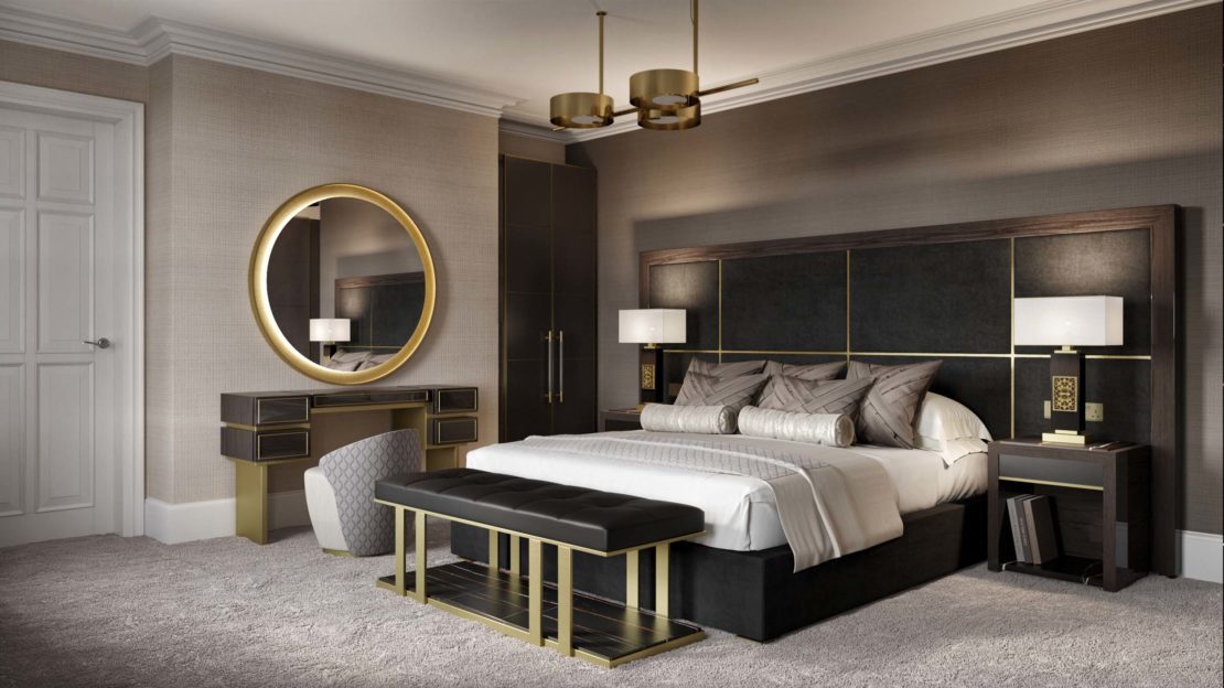 A modern bedroom in a gentleman's luxury home office features a large bed with gray and white bedding, flanked by two nightstands with lamps. A round mirror hangs above a vanity with drawers, accompanied by a cushioned stool. A bench sits at the foot of the bed. The decor includes gold accents.