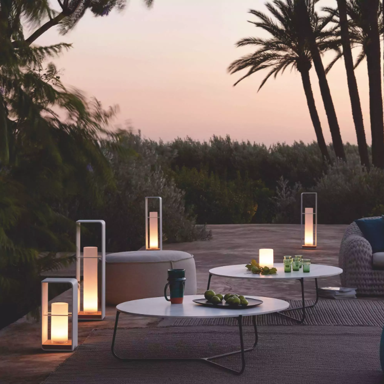 Outdoor patio with modern furniture, including circular tables holding fruit, drinks, and candles. The setting is surrounded by lush greenery and palm trees, with lanterns providing soft lighting at dusk. The ambiance is serene and inviting, perfect for a Wimbledon-style lawn party.