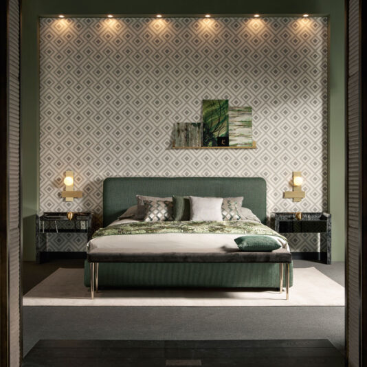 A modern bedroom with geometric wallpaper features a green cushioned headboard and a matching Contemporary Curved Upholstered Bed adorned with decorative pillows and a blanket. Two black nightstands, each topped with a yellow lamp, flank the bed. Above the headboard, two abstract wall art pieces are displayed for a contemporary touch.