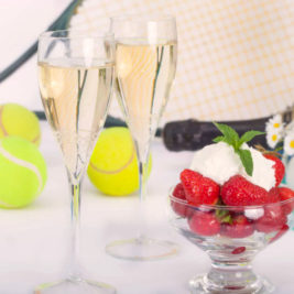 Two champagne flutes filled with sparkling wine, a glass bowl of strawberries topped with whipped cream and mint, and tennis equipment including balls and racquets in the background evoke a Wimbledon-style lawn party. Daisies are also part of the scene.