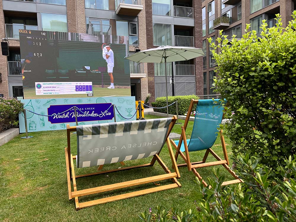 Two deck chairs and a large parasol are set up on a lawn in front of an outdoor screen displaying a tennis match. The setup exudes the charm of a Wimbledon-style lawn party within a modern residential complex, with balconies and brick facades visible in the background. A "Chelsea Creek" branded banner is also present.