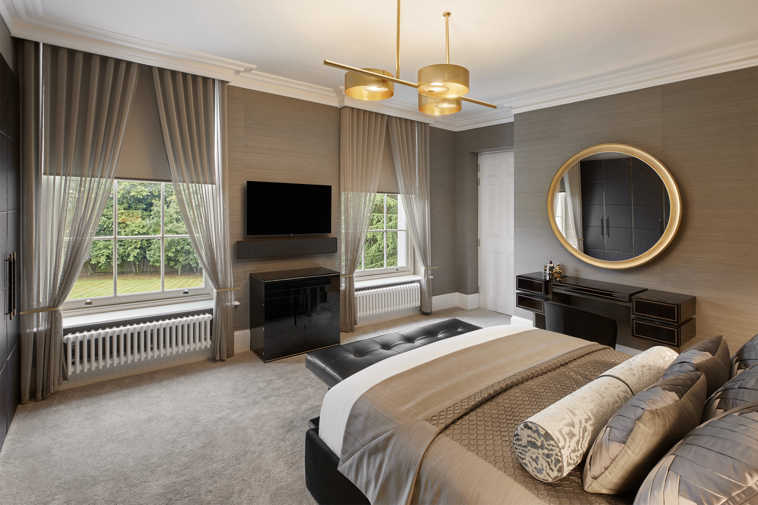 A modern bedroom with a large bed, gray and beige decor, and a round mirror above a dark wooden dresser. Two large windows with sheer curtains let in natural light. A mounted flat-screen television is centered between the windows.