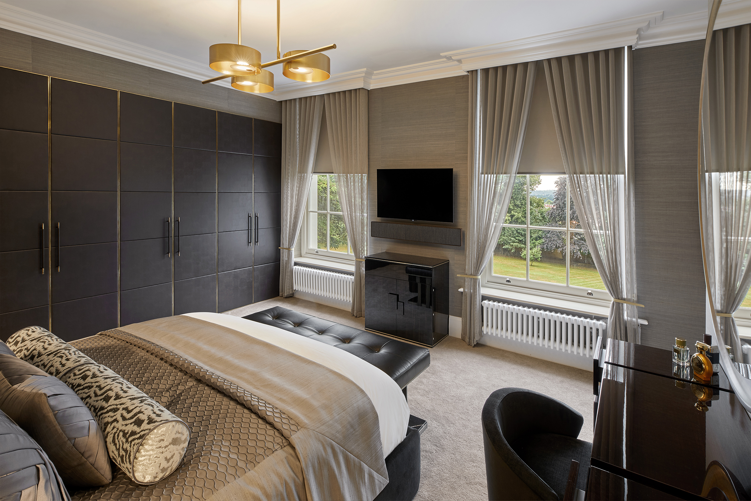 A luxurious bedroom features a bed with patterned pillows and a padded headboard, a black bench at the foot, large wardrobes, a wall-mounted TV, a desk with a chair, and tall windows with gray curtains providing a view of a lush garden outside.