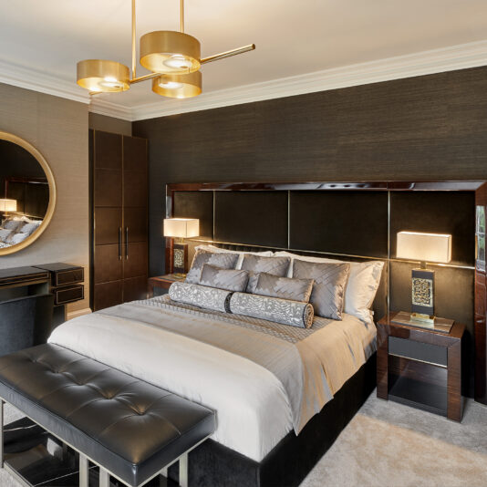 A modern bedroom featuring a large bed with grey linens and multiple pillows, flanked by nightstands with lamps. A cushioned bench sits at the foot of the bed. The room includes a dressing table with a round mirror and an upholstered chair. A sleek chandelier hangs from the ceiling.