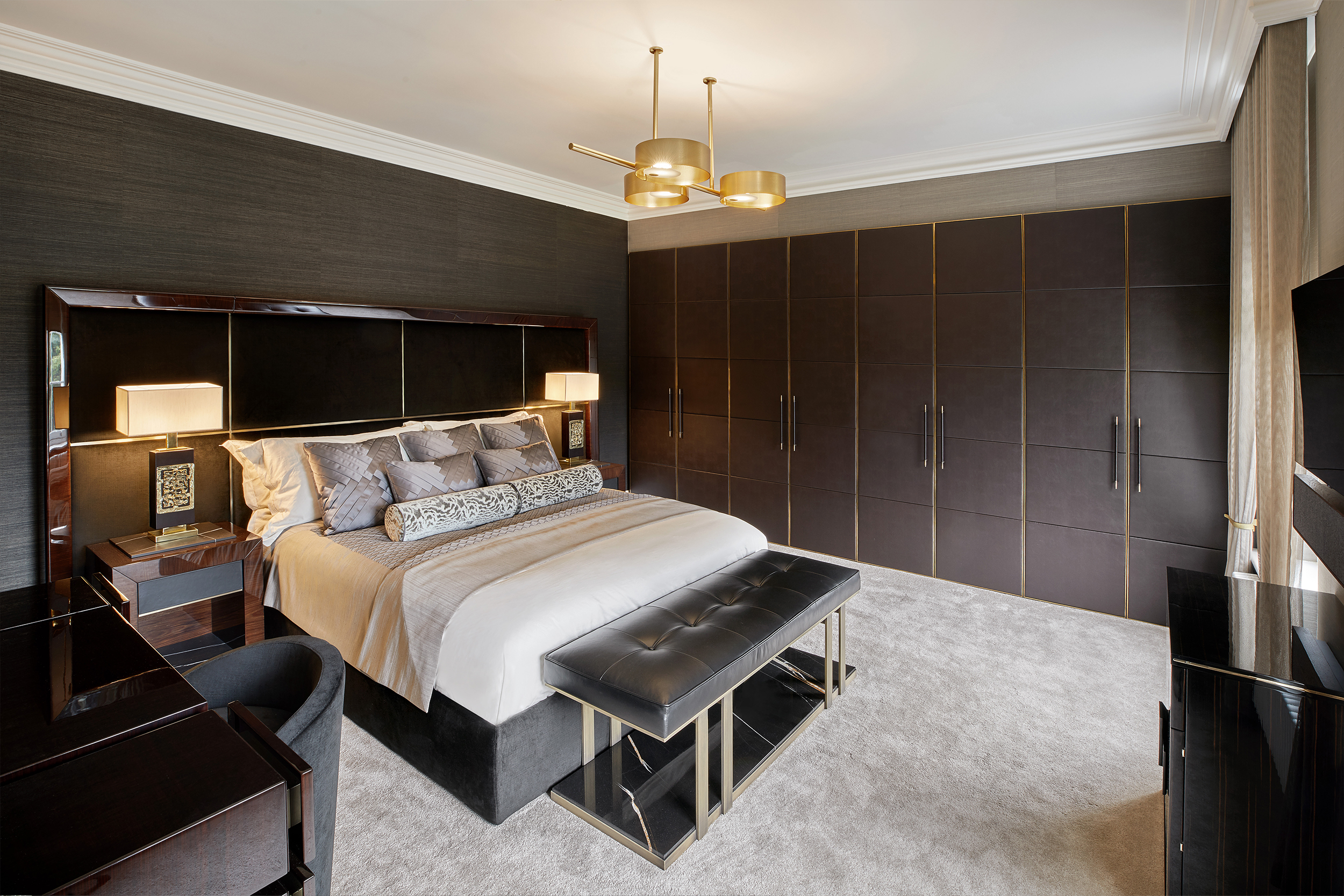 A modern bedroom features a large, dark wood bed with a neutral bedspread and multiple pillows. It includes a matching wood headboard, nightstands with lamps, a black bench at the foot of the bed, gold lighting fixtures, and a wall of dark cabinets with sleek handles.