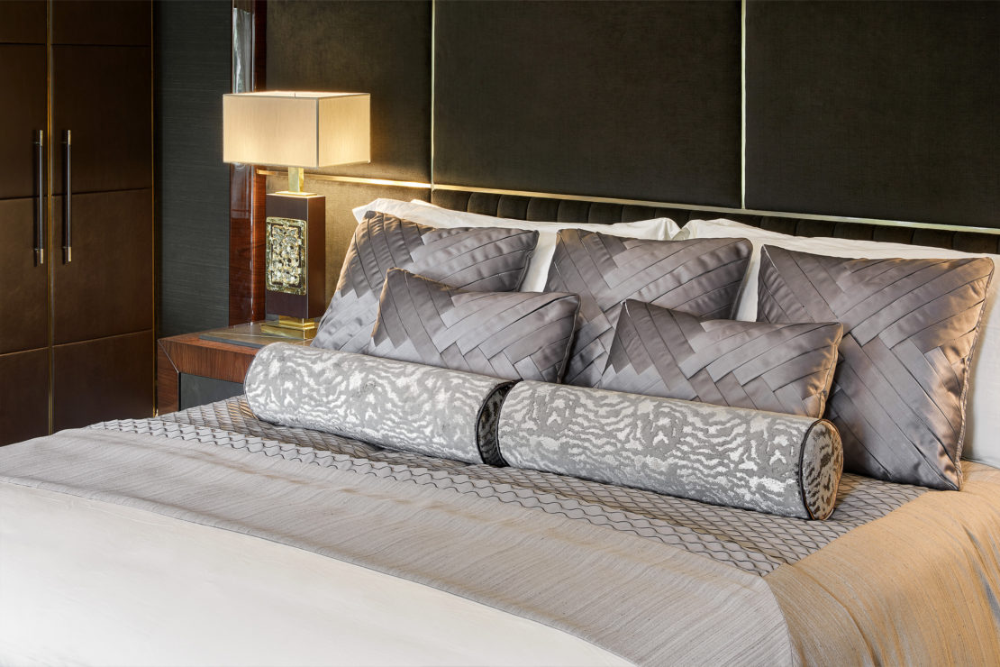 A modern bedroom with a dark tufted headboard, a neatly made bed with a light grey and white comforter, and multiple matching grey decorative pillows and bolsters. A wooden nightstand with a square lamp and a closed wardrobe are visible in the background.