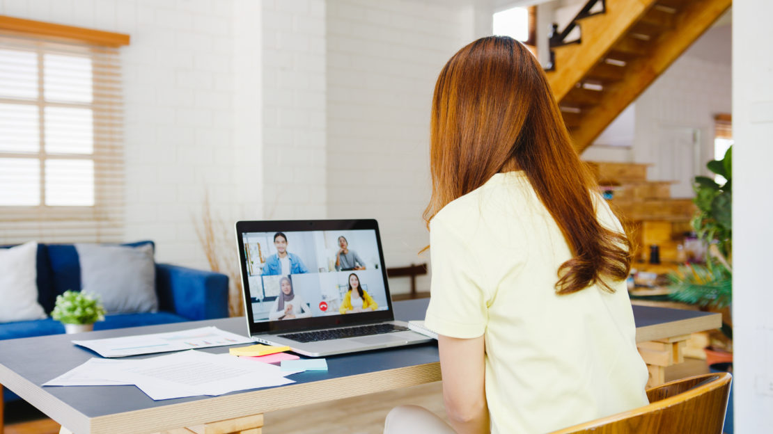 A woman with long brown hair sits at a desk in a bright, modern living room, participating in a video conference on a laptop. Papers and colorful sticky notes are spread on the desk. A staircase and blue couch are visible in the background. Learn Interior Design with us in such inspiring surroundings!