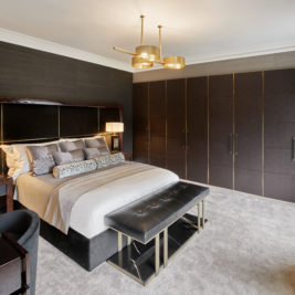 A modern bedroom features a large bed with neatly arranged pillows, flanked by two bedside tables with lamps. A bench sits at the foot of the bed. The room is designed with dark tones, including a large wardrobe along one wall and matte black furnishings.