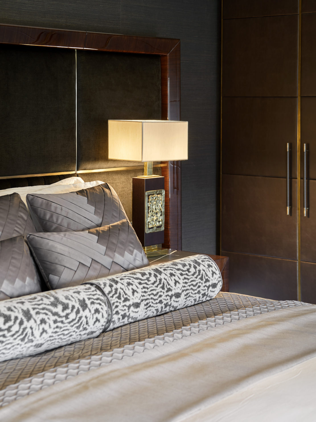 A modern, luxurious bedroom with a neatly made bed featuring grey and patterned pillows and blankets. A square, beige lampshade sits on a bedside table, illuminating the dark headboard and wooden paneling of the wall. A large, brown wardrobe stands nearby.