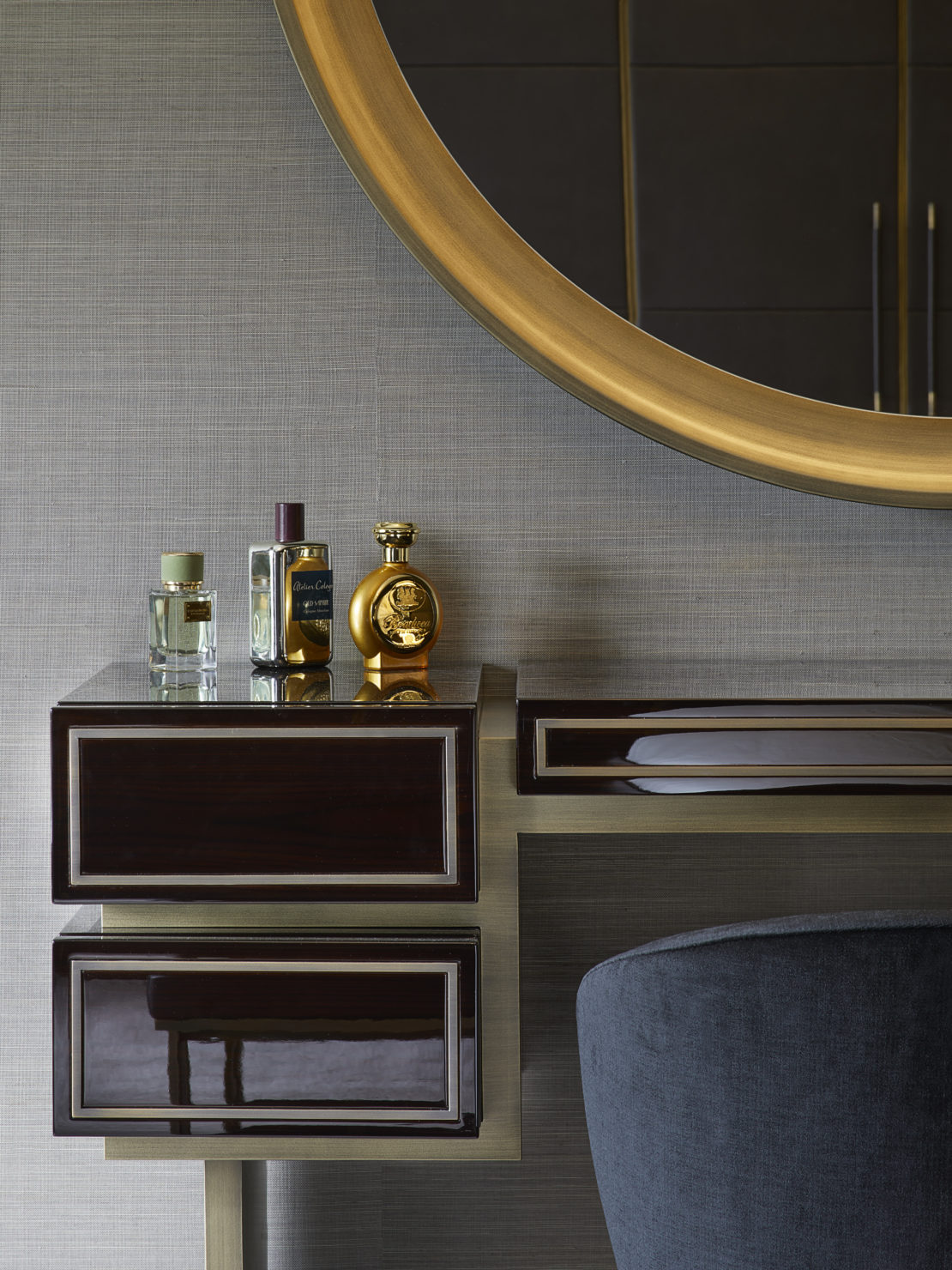 A sleek vanity with a round gold-framed mirror is adorned with a few elegant perfume bottles. The vanity features dark, reflective drawers and a gold frame. A luxurious grey chair is partially visible in the lower right corner.