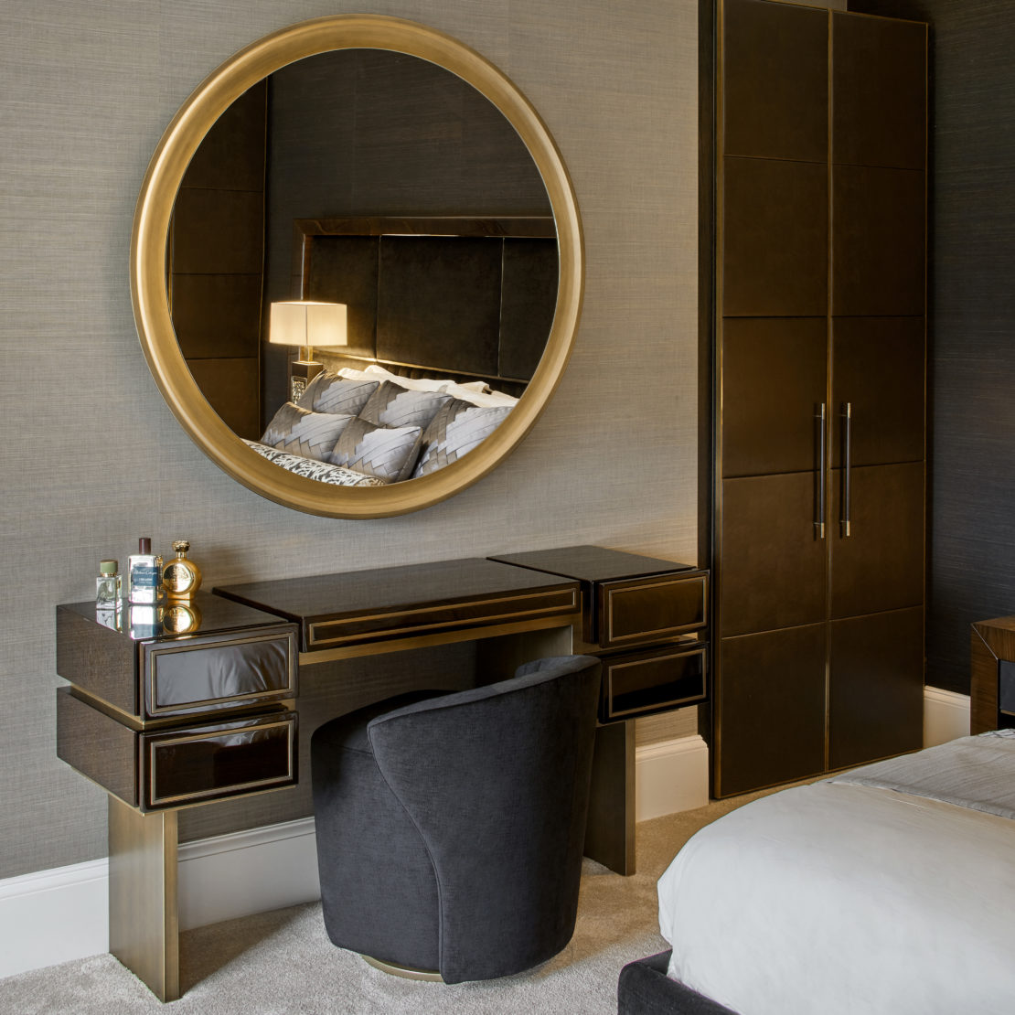 A modern bedroom exudes a blend of elegance and functionality, highlighting a gentleman's luxury home office project. It features a sleek vanity with a round gold-framed mirror, dark wood desk with drawers, and a cushioned chair. The vanity is adorned with perfume bottles, while behind it stands a tall wardrobe against a textured wall. A bed is partially visible.