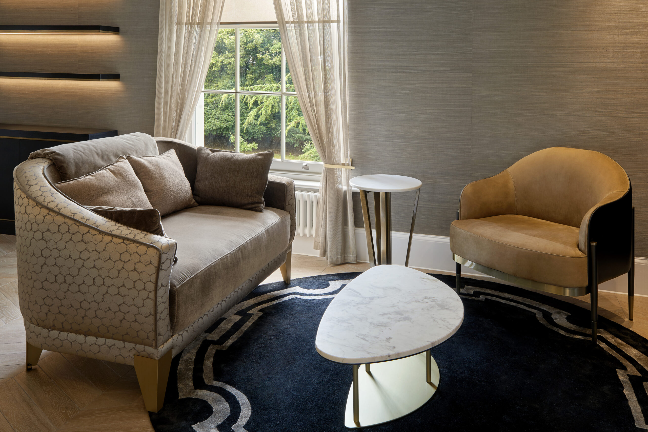 A modern living room with a beige loveseat adorned with multiple cushions, a tan armchair, and a small round white table. The room features a dark navy rug with geometric patterns and a triangular marble coffee table. A window with sheer curtains shows green foliage outside.