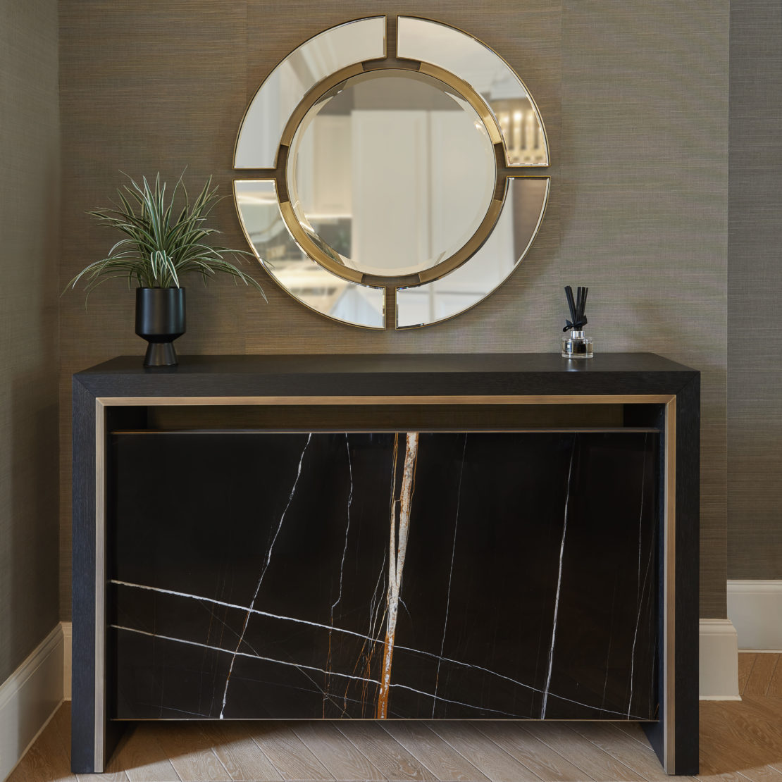 Modern console table with a black marble design, topped with a potted plant and a small decor item in a sleek room. A circular mirror with an artistic frame hangs on the wall above the table, all set against a textured wallpaper backdrop.