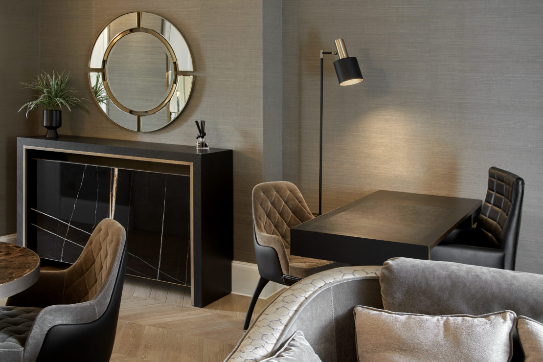 Modern living room with a wall-mounted circular mirror above a wooden console table adorned with a potted plant and a decorative item. There's a dining area with a black table, grey cushioned chairs, and a floor lamp. Two grey armchairs are partially visible in the foreground.