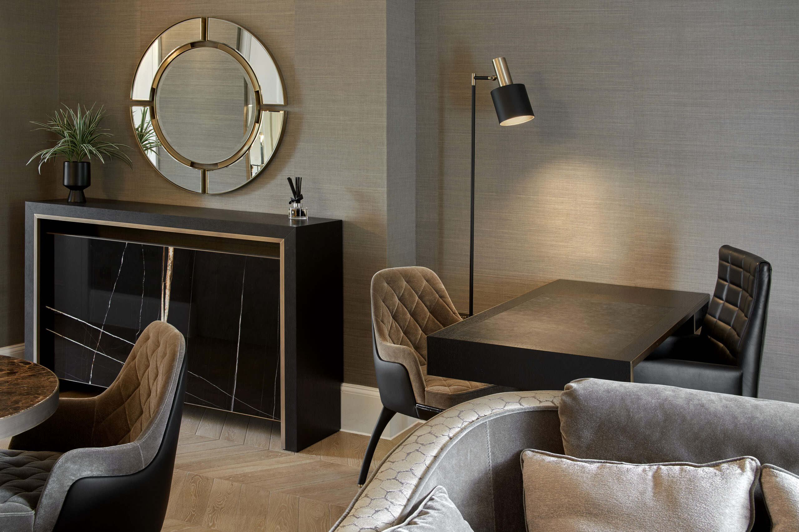 Modern living room with a wall-mounted circular mirror above a wooden console table adorned with a potted plant and a decorative item. There’s a dining area with a black table, grey cushioned chairs, and a floor lamp. Two grey armchairs are partially visible in the foreground.