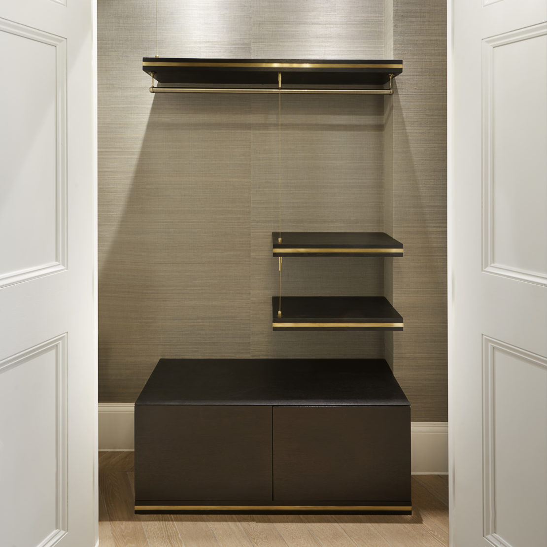 Minimalist walk-in closet with light beige walls and wooden flooring. Features include dark wooden shelves and a bench with gold accents, a hanging rod for clothes, and three small floating shelves on the right side. White double doors frame the entrance.