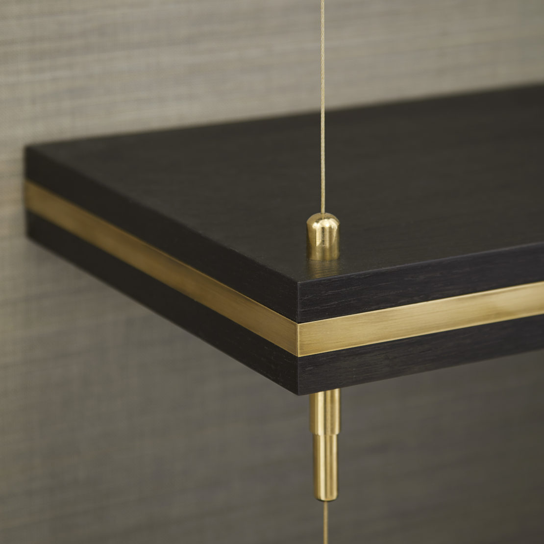 A close-up image of a dark wooden shelf with a brass accent running horizontally through the center. The shelf is supported by thin, vertically hanging brass wires. The background features a textured, neutral-colored wall.