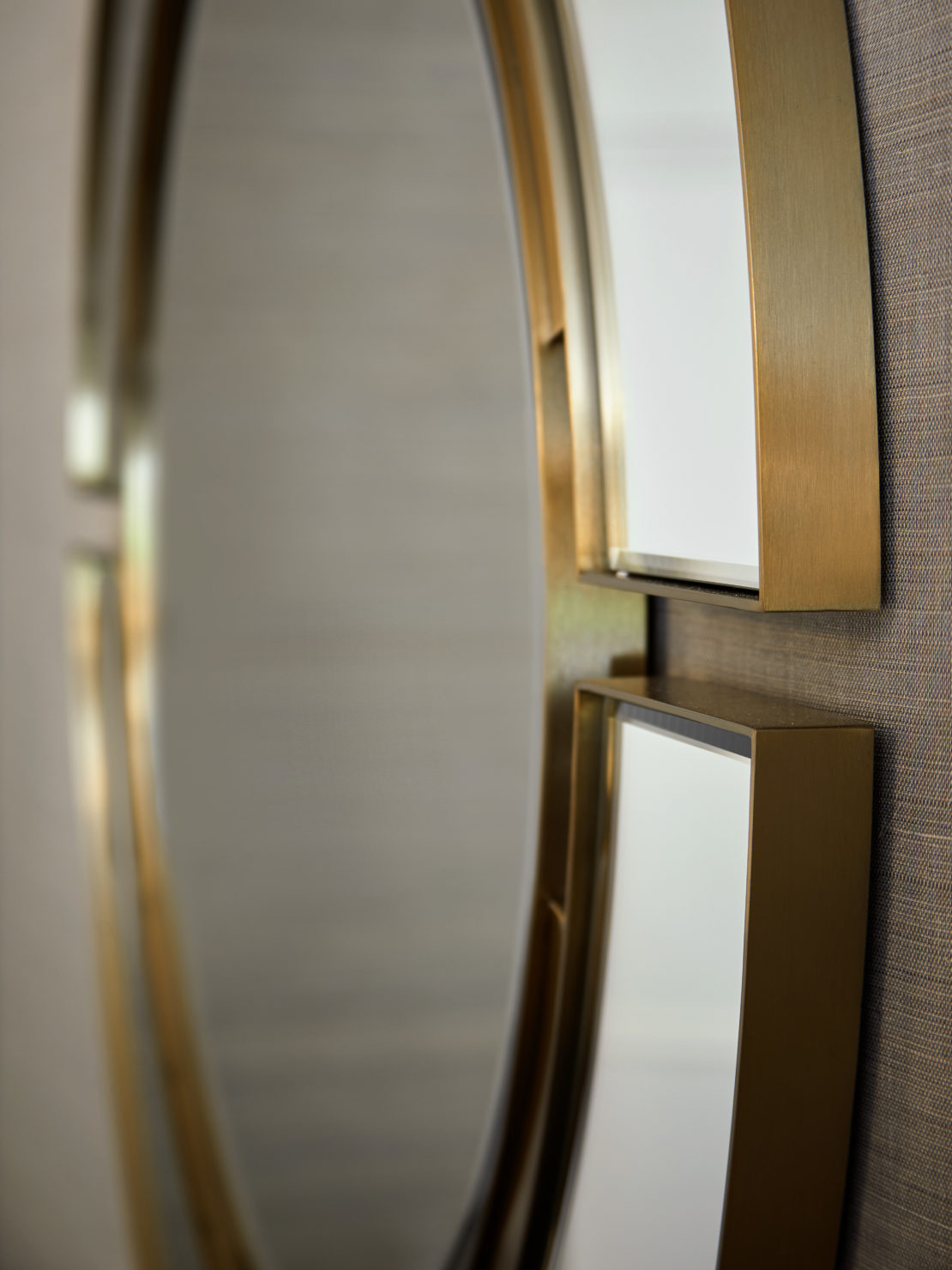 Close-up of a decorative wall mirror. The mirror features an elegant design with a circular shape and a gold-colored frame. The frame has overlapping segments, creating a modern geometric look. The background is a neutral-colored wall with a subtle texture.