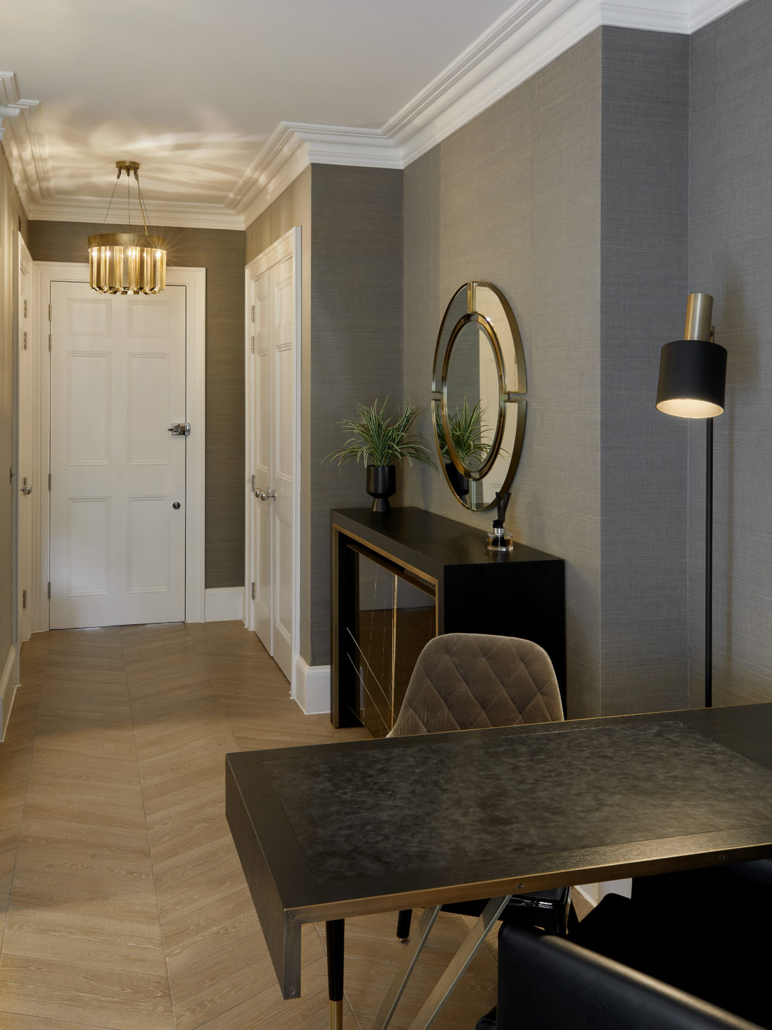 A modern hallway with greyish walls, white doors, and light wooden herringbone flooring. A black desk with gold accents and a plush chair is positioned near the entrance. A console with a mirror and potted plants is against the wall, beneath a pendant light.