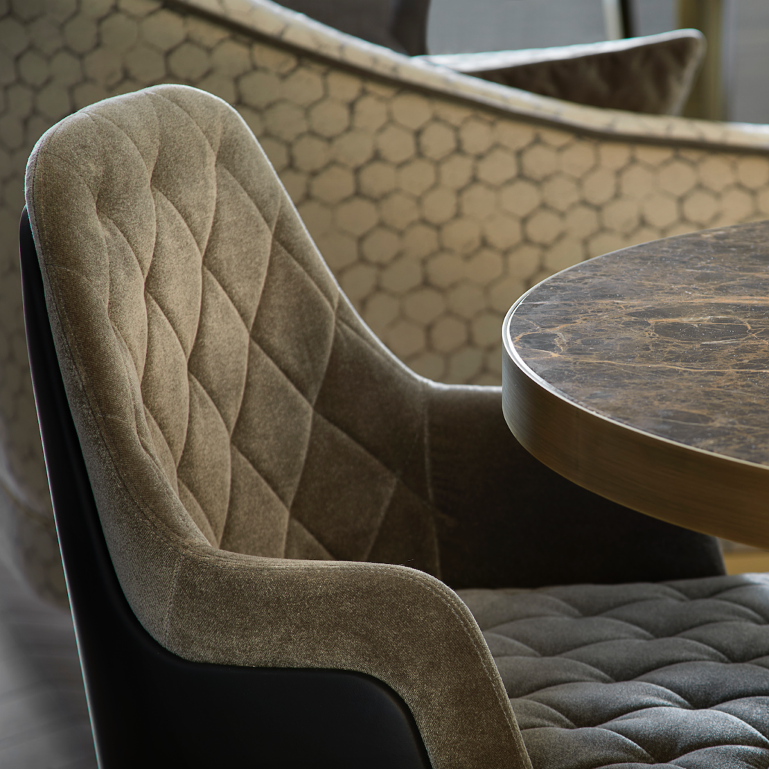 A close-up of a plush, quilted chair with a diamond pattern in greyish-brown fabric, positioned next to an elegant round table with a marble top. In the background, another chair with a hexagonal patterned fabric is partially visible.