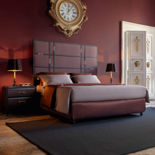 A luxurious bedroom showcases a Classic Snaffle Upholstered Bed With Tall Headboard, accompanied by two bedside tables with matching lamps. The room is enhanced by rich, deep red walls, a vintage clock above the headboard, and an ornate white door with gold accents.