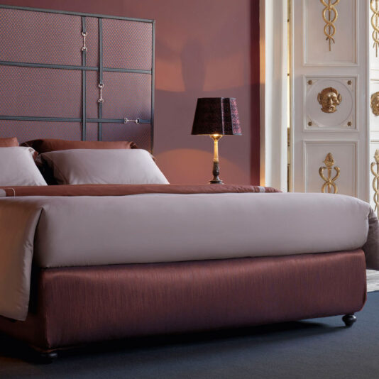 A luxurious bedroom features a Classic Snaffle Upholstered Bed With Tall Headboard, adorned with plush crimson bedding and white pillows. A bedside table with a lamp sits next to the bed. The room boasts ornate white and gold detailing on the door and a dark carpeted floor. The wall is painted maroon.