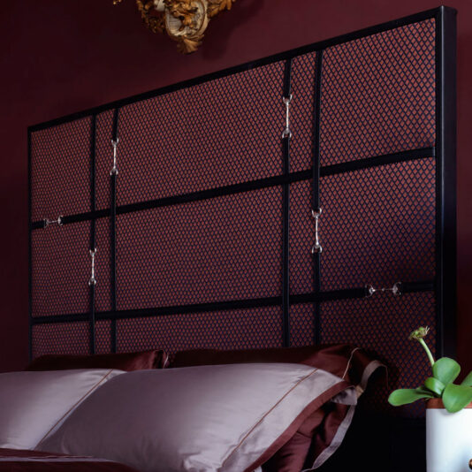 A modern bedroom features the Classic Snaffle Upholstered Bed With Tall Headboard, adorned with distinctive hooks. The wall behind the bed is painted a rich burgundy, while the bedding includes pillows and sheets in various shades of brown. A potted plant on a nearby surface adds a touch of nature to the space.