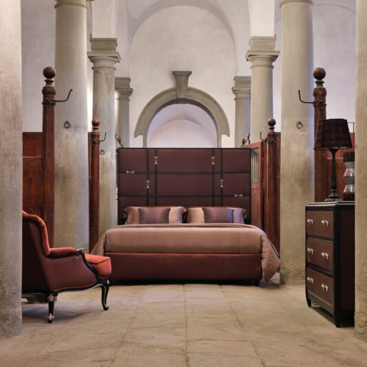 A stylish bedroom features the Classic Snaffle Upholstered Bed With Tall Headboard, paired with matching pillows and duvet, set against tall, arched columns. To the left sits a maroon armchair, while to the right stands a dark wooden dresser topped with a decorative lamp. The room is accented by a rustic stone floor.