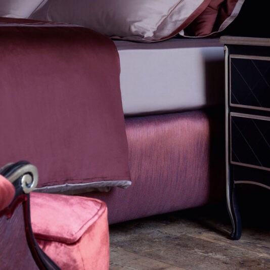 A cozy bedroom showcases the Classic Snaffle Upholstered Bed With Tall Headboard, adorned with mauve sheets. Beside the bed stands a black nightstand featuring a diamond pattern, while a plush pink armchair is partially visible in the foreground.