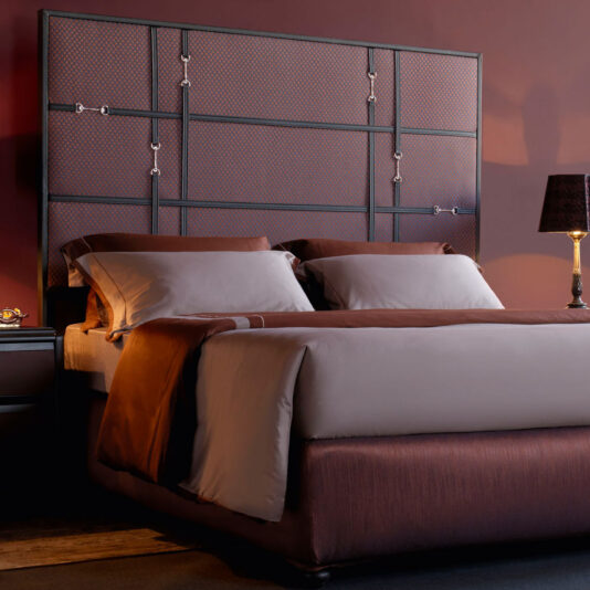 A modern bedroom with a maroon color scheme features the Classic Snaffle Upholstered Bed With Tall Headboard, showcasing its grid-patterned design and metallic accents. Adorned with beige and brown bedding, the bed is complemented by a bedside table on the right side, hosting a lit table lamp with a black shade.