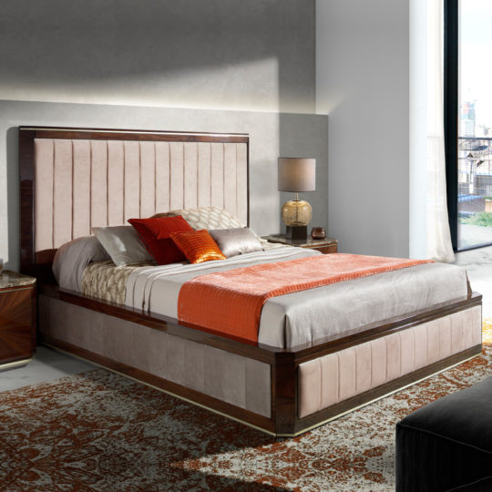 A modern guest bedroom with a large bed featuring an upholstered headboard and frame, dressed in neutral-colored bedding with orange accents. To the right, there's a large window with cityscape views. A nightstand with a lamp and books sits to the left of the bed.