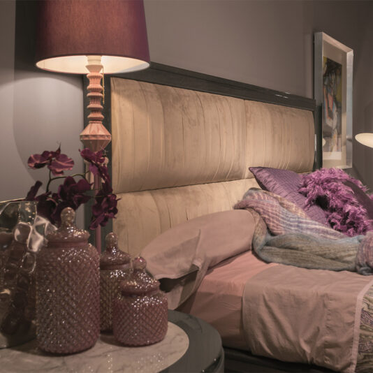 Luxury Modern Upholstered Bed A cozy bedroom features the Luxury Modern Upholstered Bed with beige fabric, adorned with pink and purple bedding. A matching lamp with a pink shade and ceramic jars sit on the bedside table. Purple flowers add a touch of decoration. The room is softly lit, exuding a tranquil ambiance.
