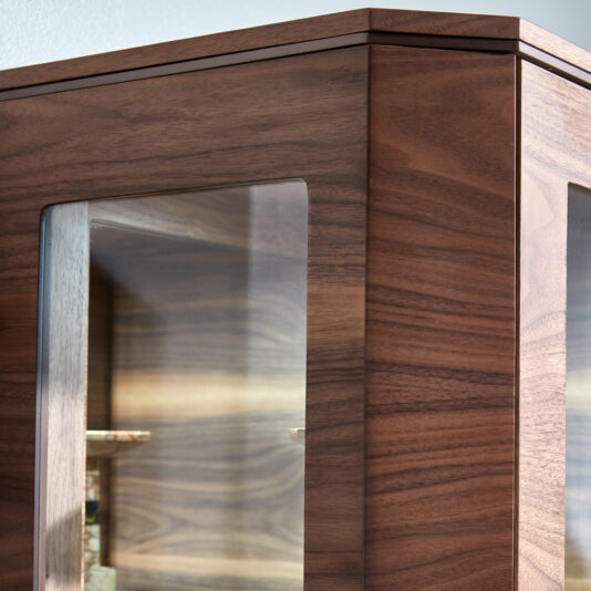 Luxury Modern Bookcase With Storage Close-up of the Luxury Modern Bookcase With Storage, featuring glass panel doors. The wood boasts a rich, dark finish, and the glass panels reveal a glimpse of the wooden interior shelves. The cabinet's corner is prominently featured in the image.