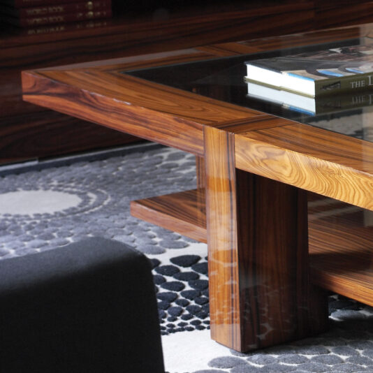 The Modern Rectangular Coffee Table with Glass Top is positioned on a dark, textured rug featuring circular patterns. Several books are neatly stacked on the table. The corner of a black sofa is partially visible in the foreground, and a wooden bookshelf can be seen in the background.