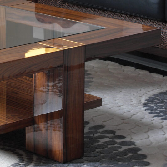 A close-up view of the Modern Rectangular Coffee Table With Glass Top showcases its polished dark wood finish and convenient lower shelf. It sits elegantly on a gray and white patterned area rug, with the edge of a black leather couch visible in the background.