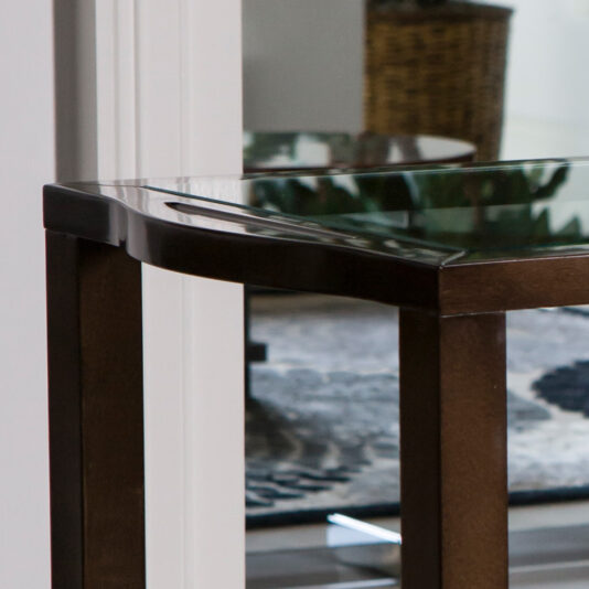 Modern Trolley With Glass Top A close-up view of the Modern Trolley With Glass Top, featuring a sleek dark wooden frame, situated in an indoor space. The background reveals a softly blurred room with a patterned rug and a wicker basket.
