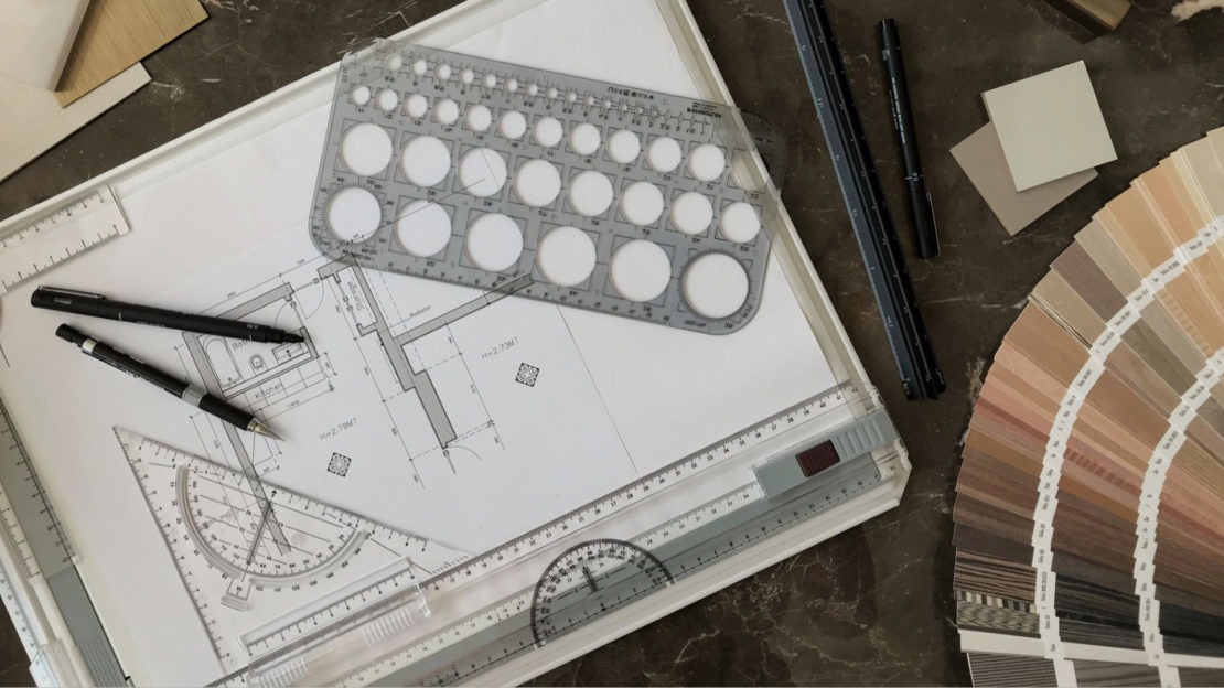 A workspace with architectural plans, drafting tools like rulers and circle templates, black pens, color samples, and a fan deck of color swatches is shown. The items are neatly arranged on a dark table alongside luxury furniture pieces, suggesting a design or drafting project in progress.