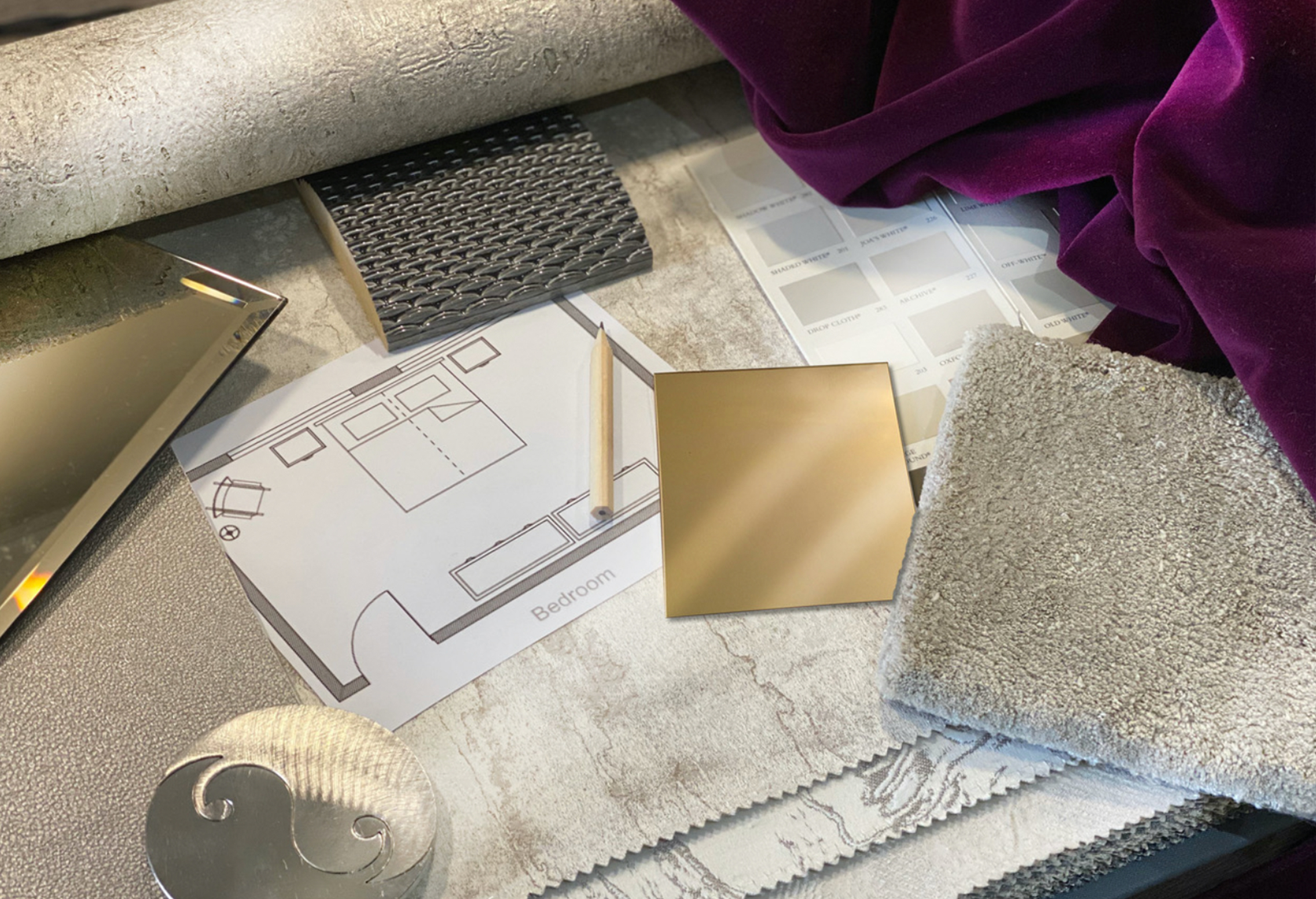 A flat lay of interior design course materials includes a floor plan, fabric swatches, textured samples, a metallic circular object, rectangular gold and mirrored samples, and a gray woven material, all placed on a table with a purple velvet cloth draped nearby.