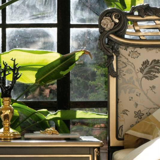 Ornate Upholstered Italian Bed An Ornate Upholstered Italian Bed featuring a patterned headboard is positioned beside a window. Large green leaves of indoor plants are visible through the glass. On the nightstand beside the bed are a unique black tree-shaped sculpture and a golden lamp.
