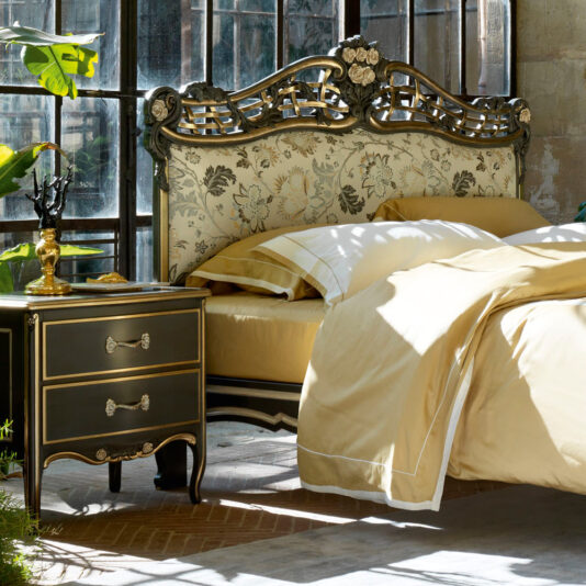Ornate Upholstered Italian Bed A luxurious bedroom features the Ornate Upholstered Italian Bed, characterized by its intricate floral-patterned headboard and elegant gold trim. The bed is dressed in beige and white bedding. Beside it, there's a matching nightstand adorned with a golden candle holder and decorative items. Large windows bring in natural light.