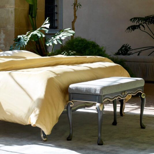 Ornate Upholstered Italian Bed A sunny bedroom featuring the Ornate Upholstered Italian Bed adorned with light yellow bedding and a grey bench at its foot. The room is accentuated by large green plants, a window with light streaming in, and walls in neutral tones, creating a serene and inviting atmosphere.