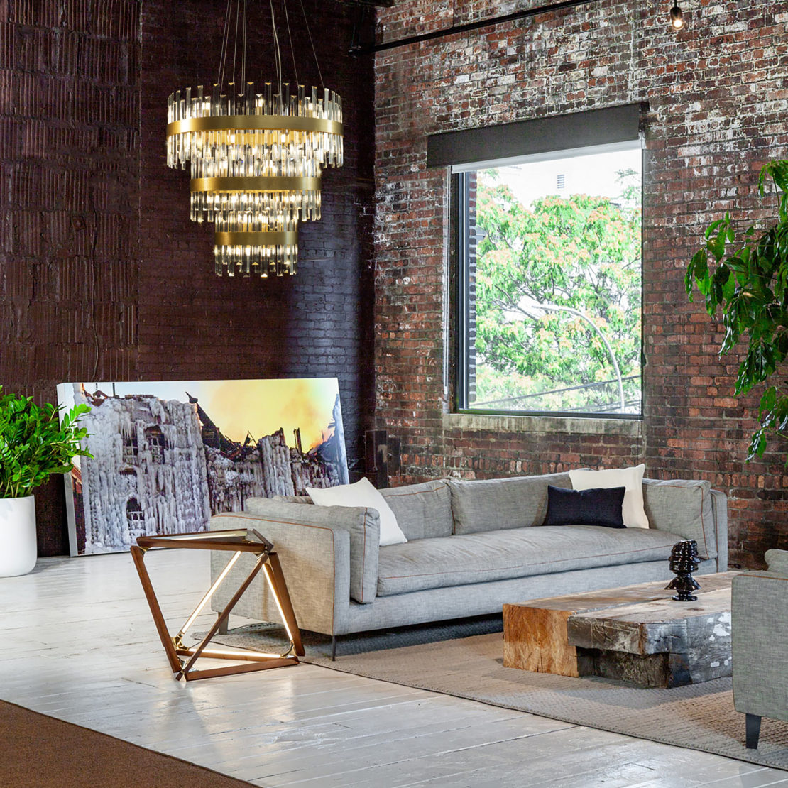 A modern living room with a gray couch adorned with white and black pillows, a geometric side table, and a wooden coffee table. A large window offers a tree view while affordable luxury lighting in the form of a chandelier hangs from the dark brick wall. A framed photograph leans against the wall.