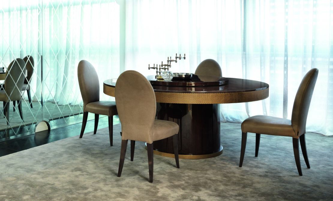 A modern dining room set with a dark round table and four beige upholstered chairs. The table features gold accents and a decorative candelabra. The dining room is lit by natural light from large windows with sheer white curtains, while the floor is covered by a neutral carpet.