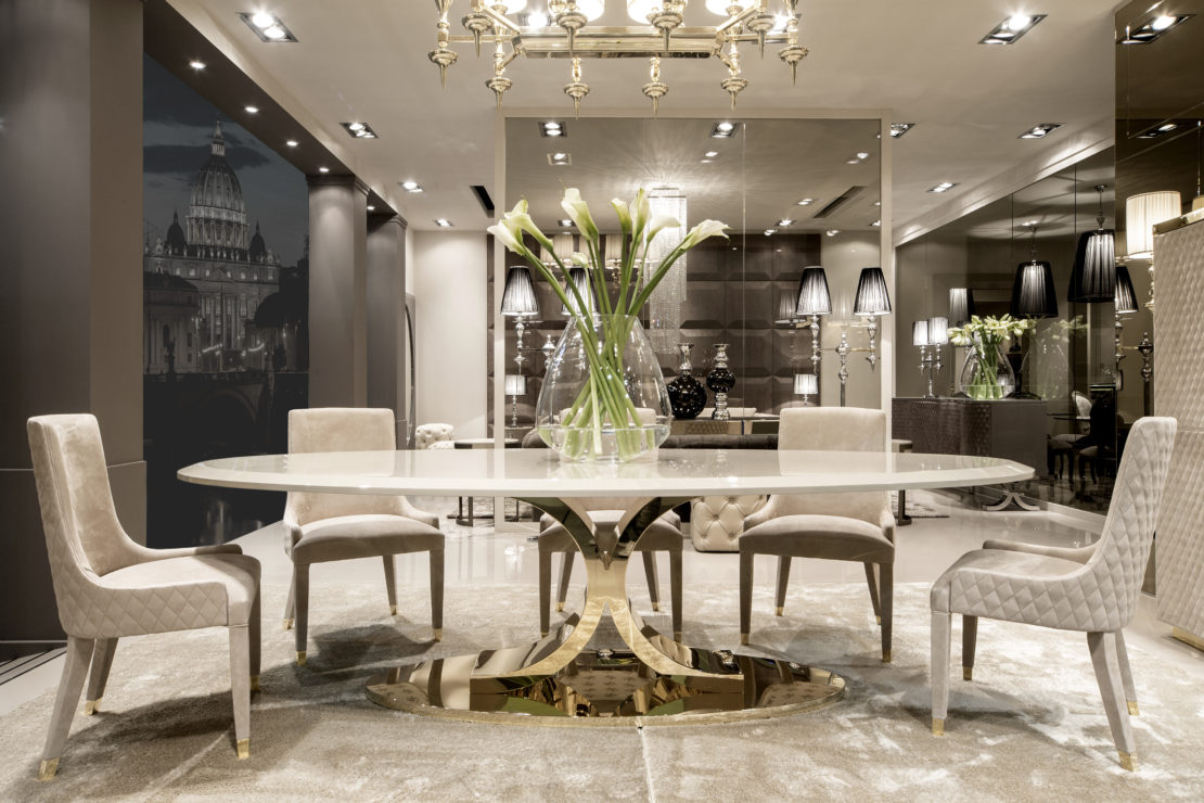 Elegant dining room with a large oval table and six plush chairs. The table is adorned with a tall vase holding white flowers. A gold chandelier hangs from the ceiling, and the dining room features modern lighting, reflective surfaces, and neutral tones.