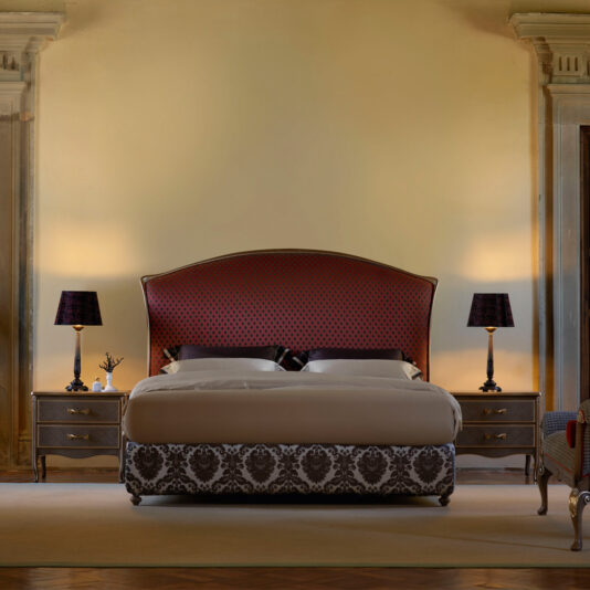 Classic Italian Bed A luxurious bedroom showcases the Classic Italian Bed, adorned with a plush, red and black patterned headboard and an ornate black and white bedspread. Flanking the bed are two wooden nightstands each topped with sleek black lamps. The room exudes elegance with its soft lighting and refined decor.