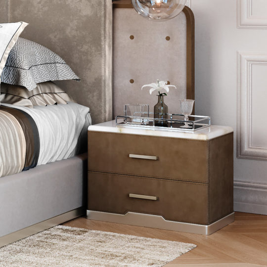 A modern bedside table with two drawers and sleek handles sits beside a stylishly made bed with gray and white bedding. A silver tray holding a flower vase and glass decanter is placed on the marble-topped table, and a round glass pendant light hangs above.