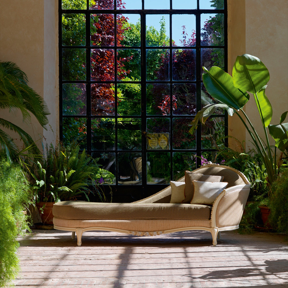 A Large Classic Louis Style Chaise Longue with cushions is set in a sunlit room with expansive grid-like windows that overlook a lush garden. The lounge is surrounded by large potted plants, creating a peaceful and inviting atmosphere.